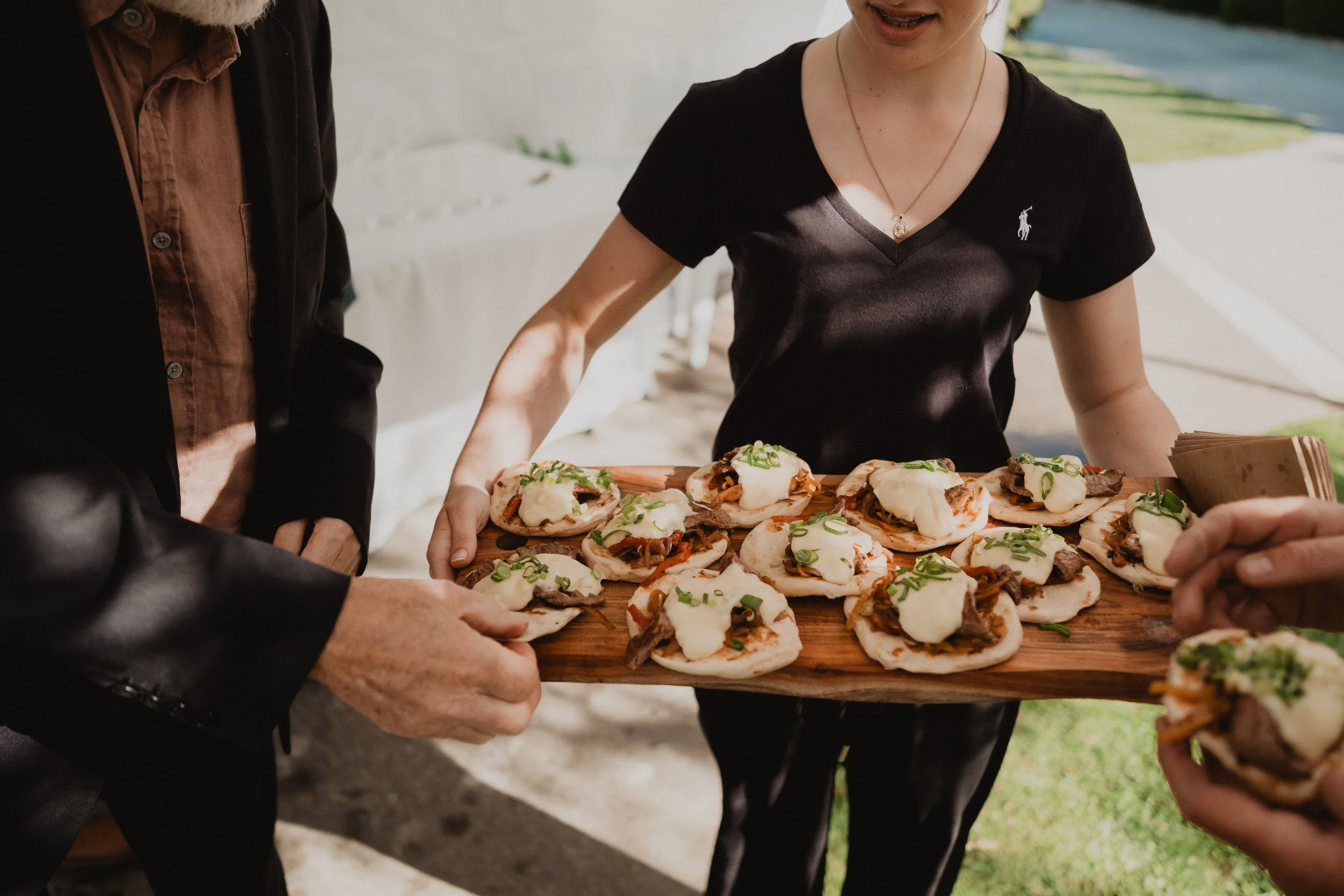 Several small tacos topped with cheese and green onions on a wooden board being offered at an outdoor event, with people reaching for them.