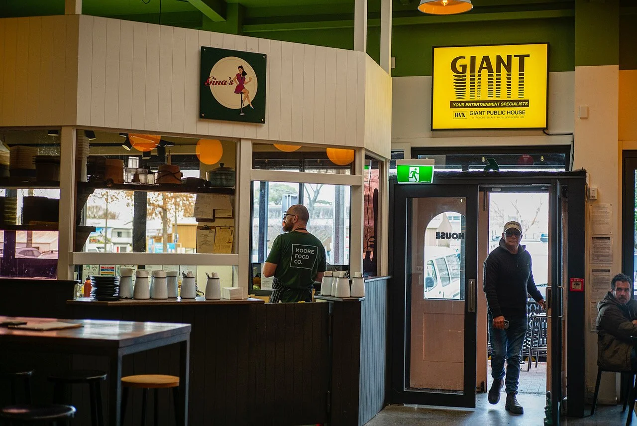Inside a restaurant with a man walking out the door wearing a hoodie and cap, another man behind the counter, and a seated man on the right. Signage includes a yellow sign reading 'GIANT' and a smaller green emergency exit sign.