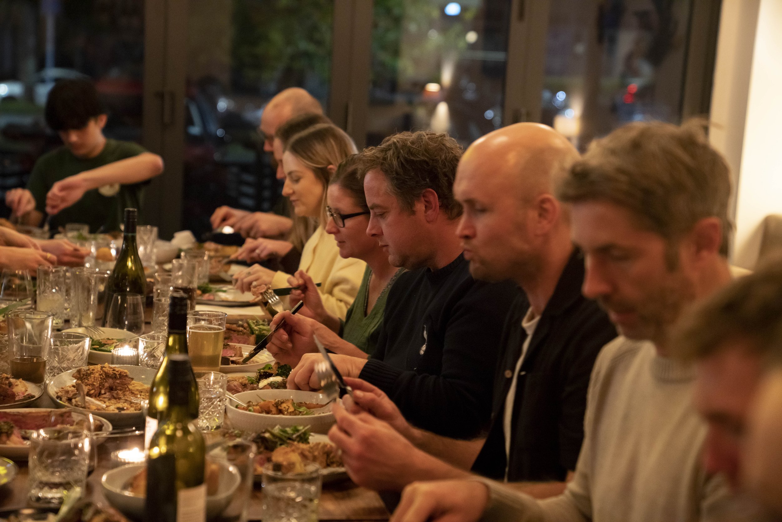 People sitting at a long dinner table enjoying a meal, with plates of food, drinks, and wine bottles in a warmly lit restaurant or home setting.