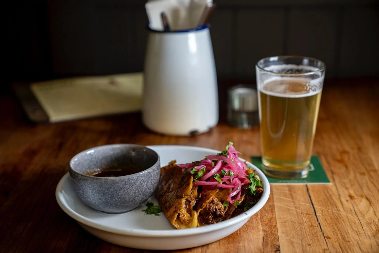Plate with a taco topped with pink pickled onions and herbs, a bowl of sauce, and a glass of beer on a wooden table.