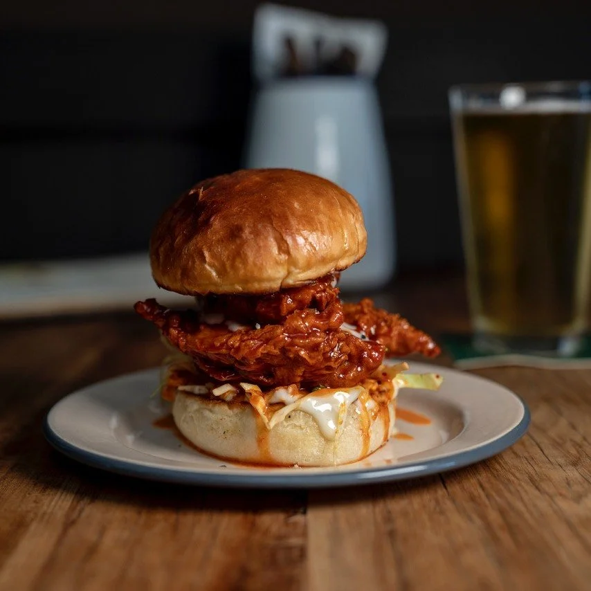 Fried chicken sandwich with coleslaw and spicy sauce in a brioche bun, served on a white plate with a drink in the background.