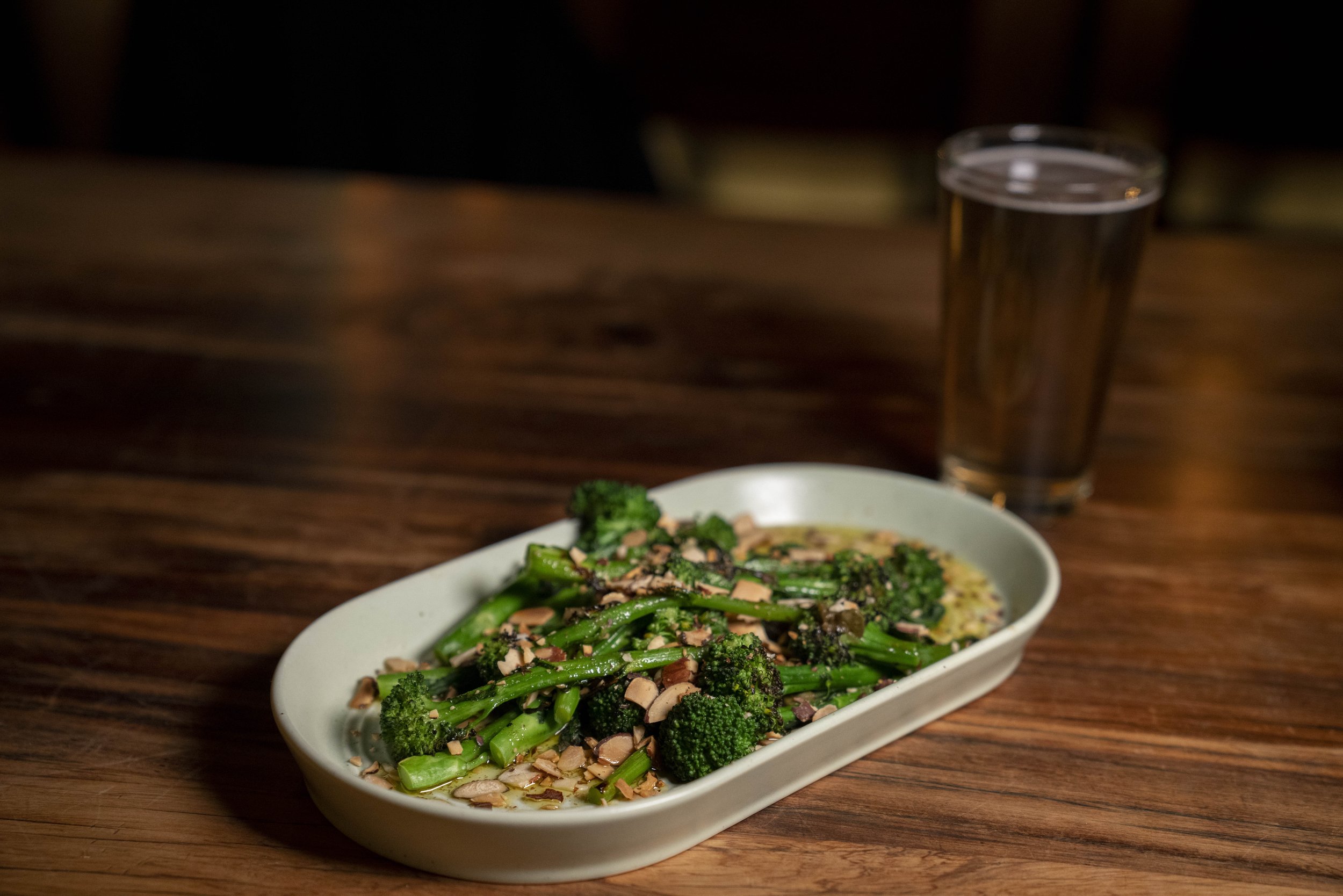 A white salad bowl filled with sautéed broccoli and garlic, topped with chopped almonds, sitting on a wooden table. In the background, a glass of water is visible.
