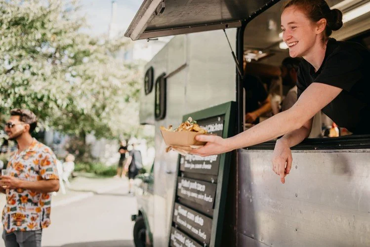 A smiling woman is serving food from a food truck, handing a bowl of chips or fries to a customer outside on a sunny day. The food truck is silver with a black menu board displayed outside.