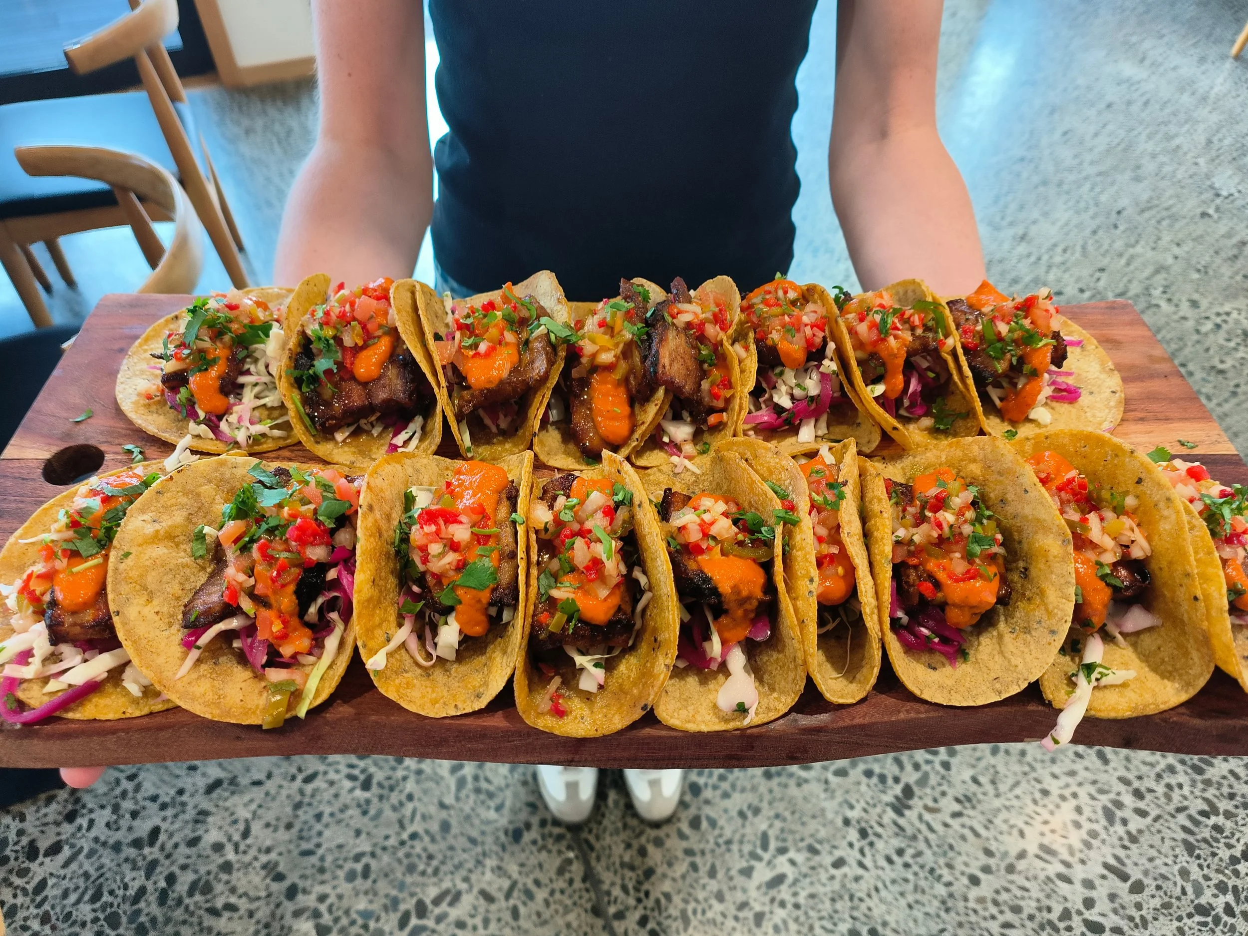 A person holding a wooden serving board with a row of tacos filled with meat, vegetables, and garnishes.