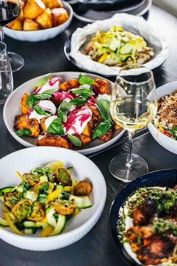 A table with various dishes including roasted vegetables with herbs, a bowl of tacos topped with shredded cheese and cucumber slices, a bowl of mixed grilled chicken and vegetables, a bowl of meat with sauce, and a glass of white wine.