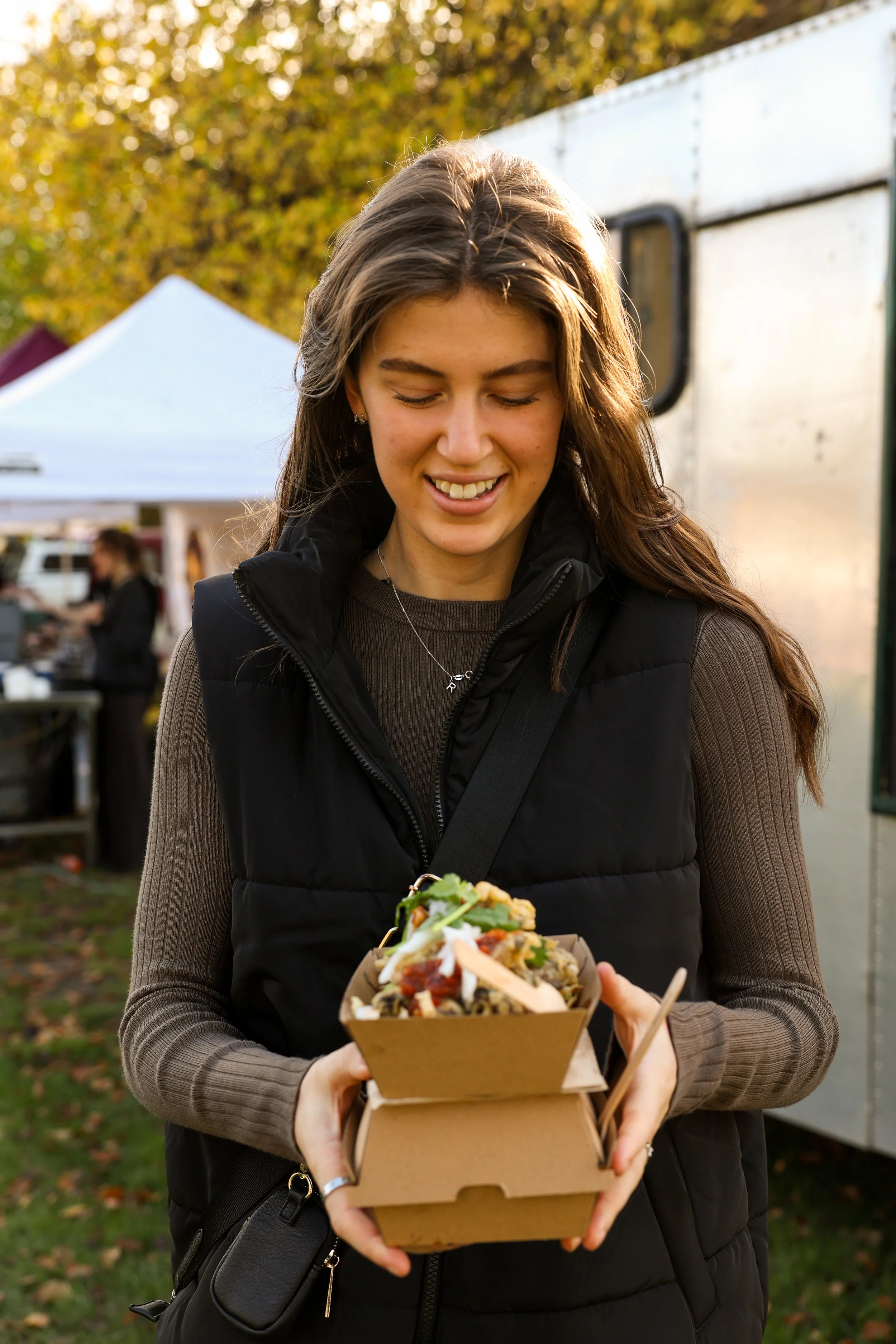 A young woman with long brown hair smiling as she receives a box of food at an outdoor event during autumn, with trees and tents in the background.