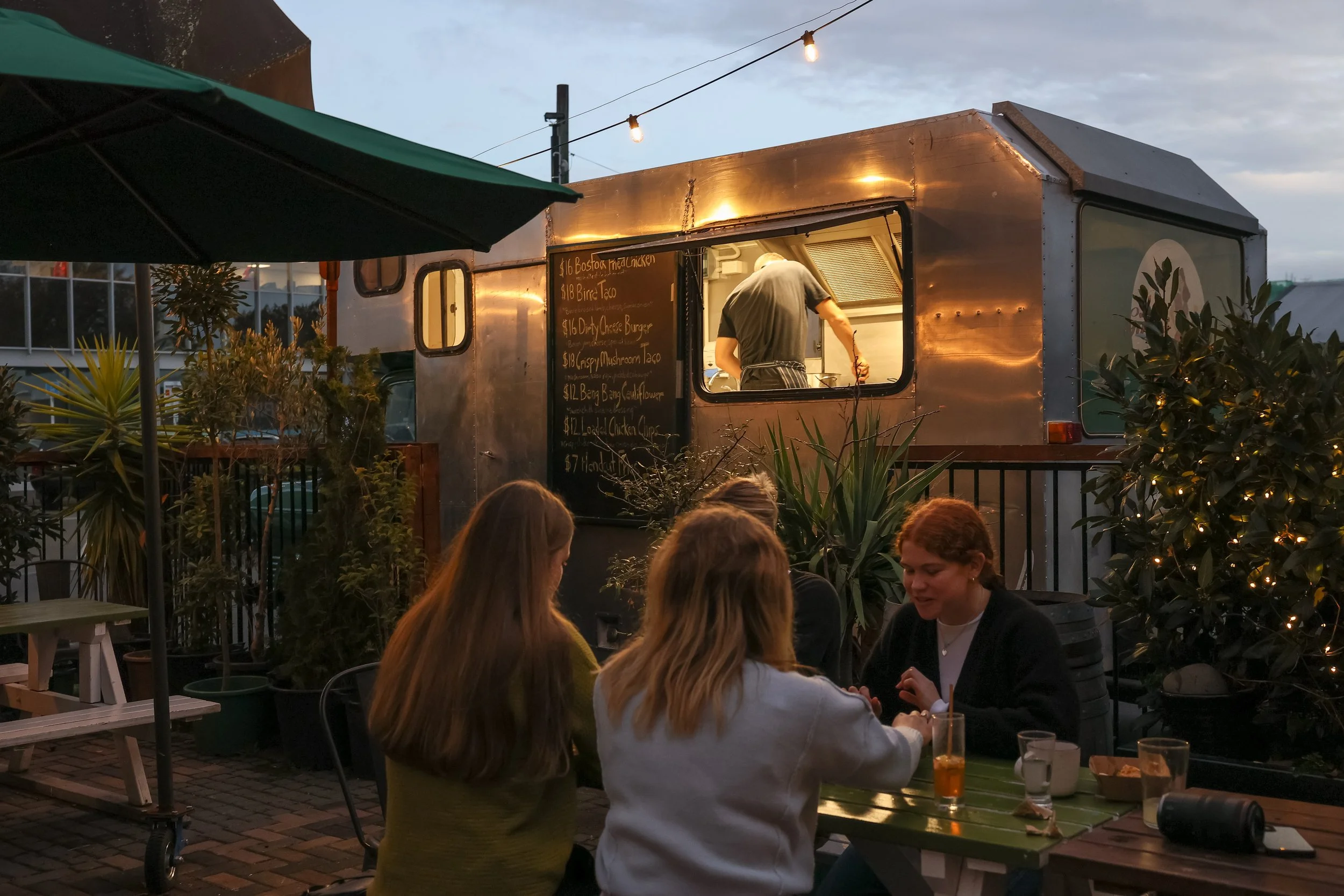People sitting at an outdoor dining table near a food truck at dusk, with string lights overhead and plants around the area.