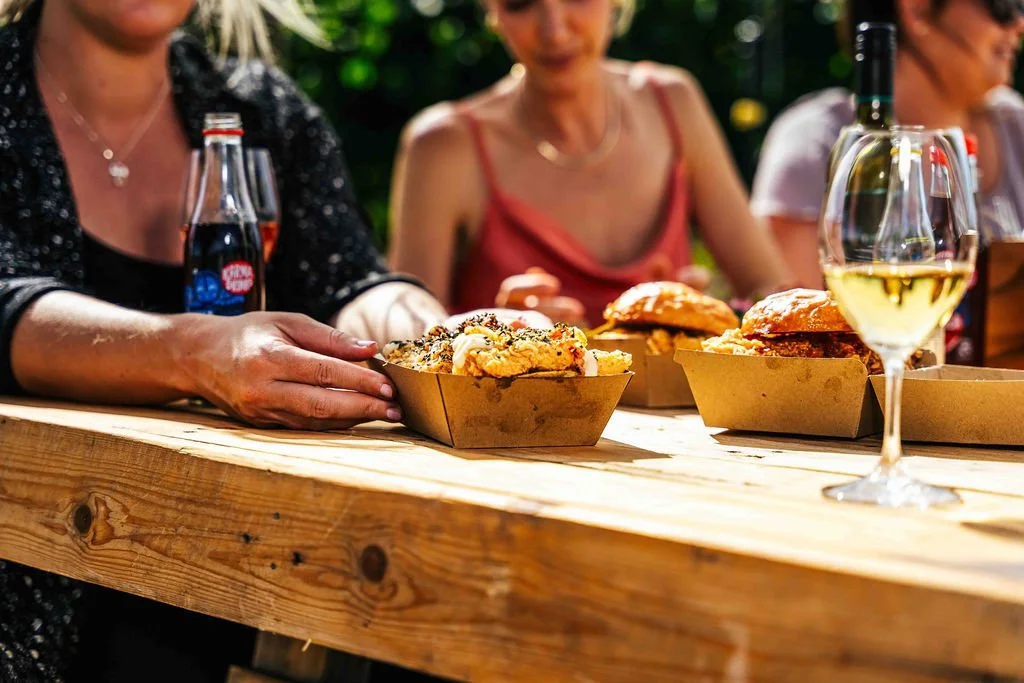People enjoying food and drinks outdoors, with sandwiches, a glass of white wine, and soda bottles on a wooden table.