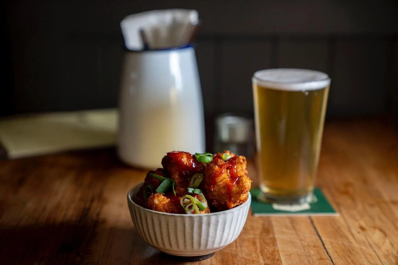 A bowl of spicy fried chicken bites garnished with chopped green onions, placed on a wooden table with a glass of beer and a salt shaker in the background.