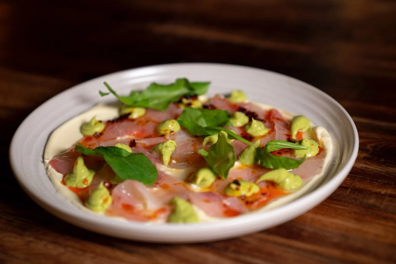 Pizza with prosciutto, green leaves, and a drizzle of green sauce on a white plate, placed on a wooden table.