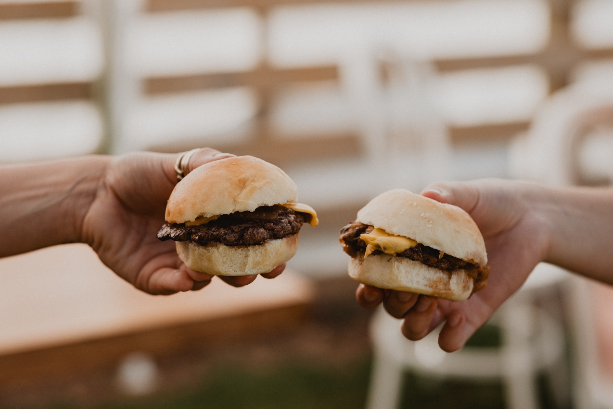 Two hands are holding small sandwiches with beef patties and cheese. The background is blurred with a light-colored wall.