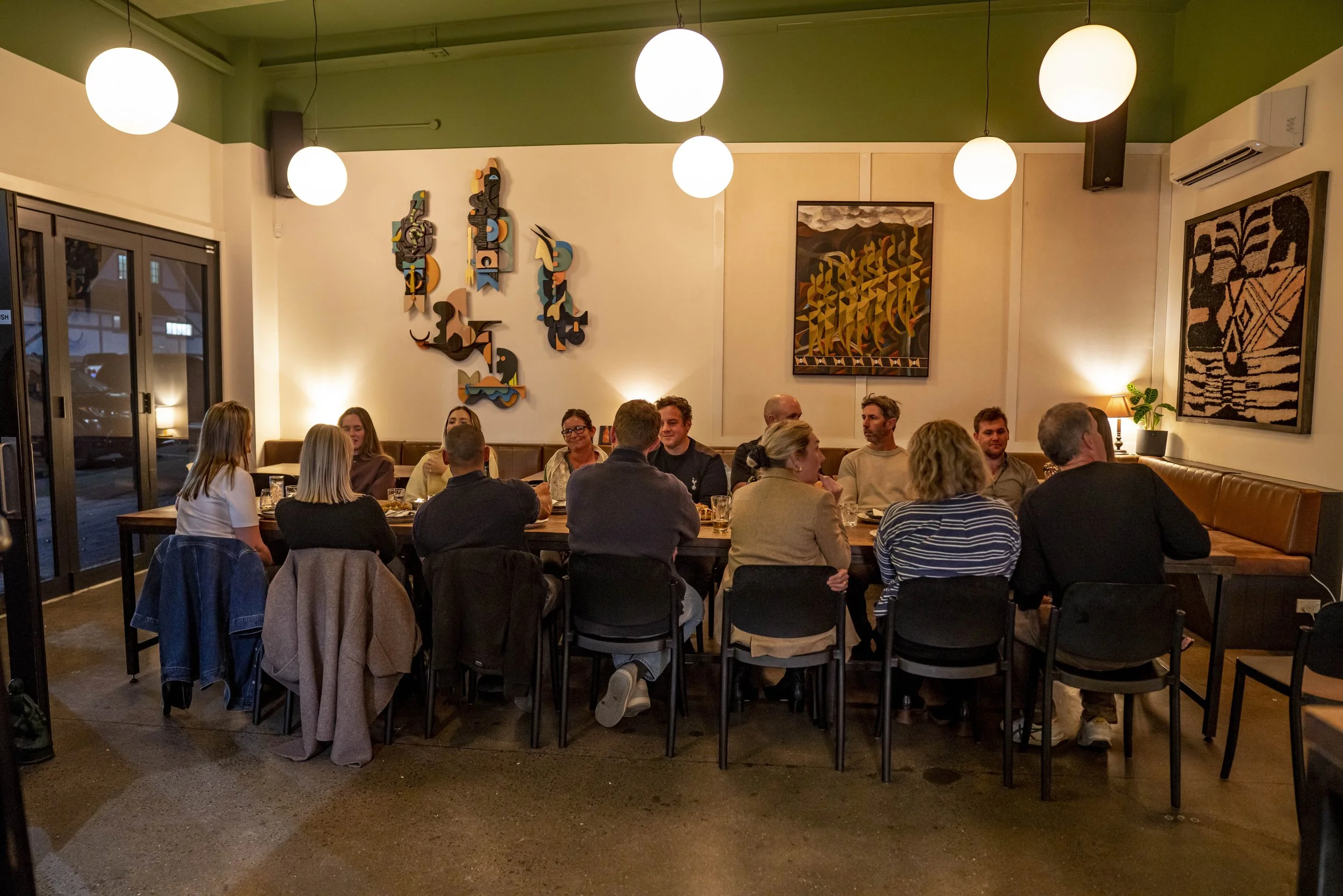 People sitting at a long table in a cozy restaurant with vibrant artwork on the walls, warm lighting, and a large window showing parked cars outside.