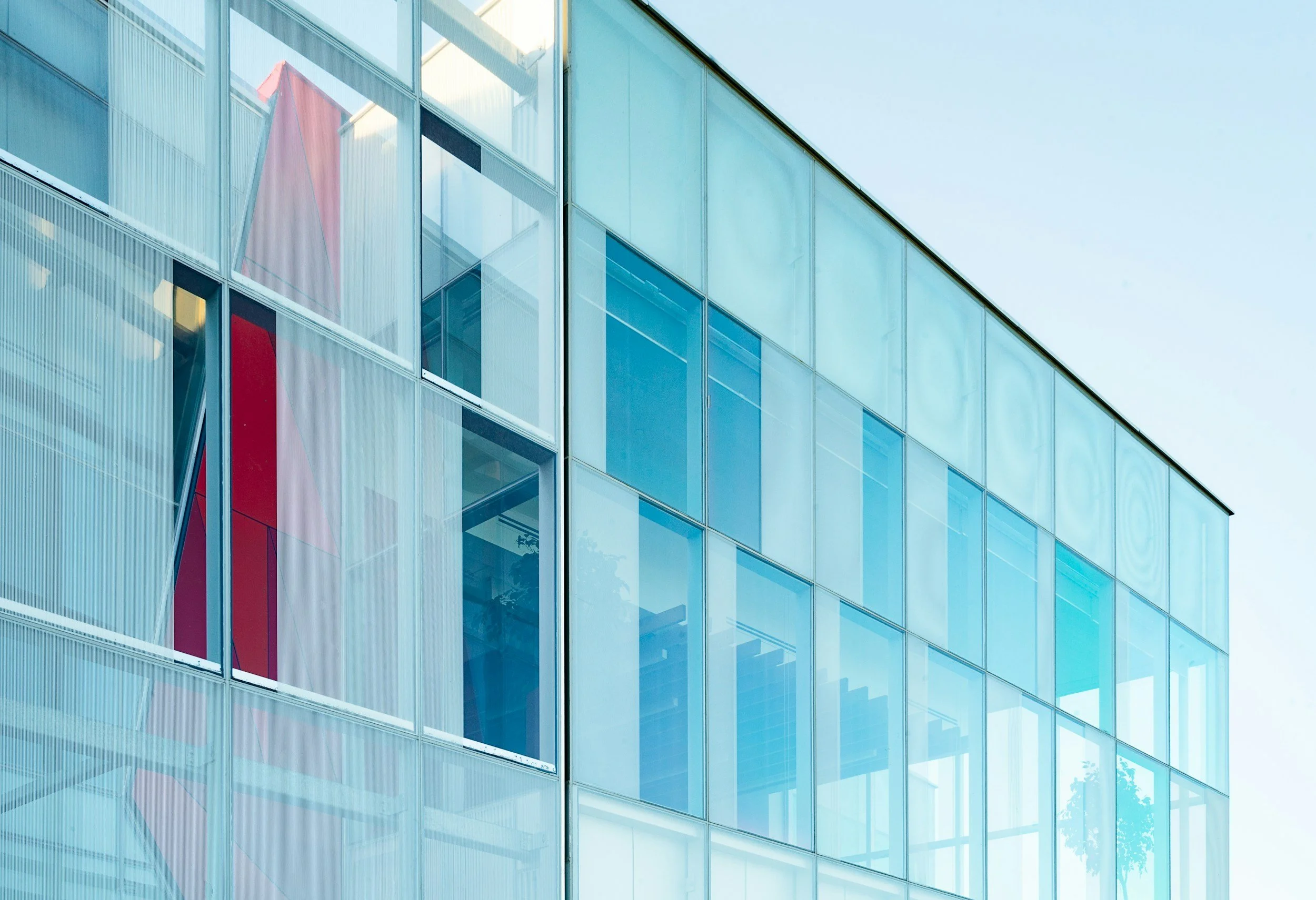Fachada de un edificio moderno con ventanas de vidrio que reflejan el cielo azul y estructuras internas, en un estilo arquitectónico contemporáneo.