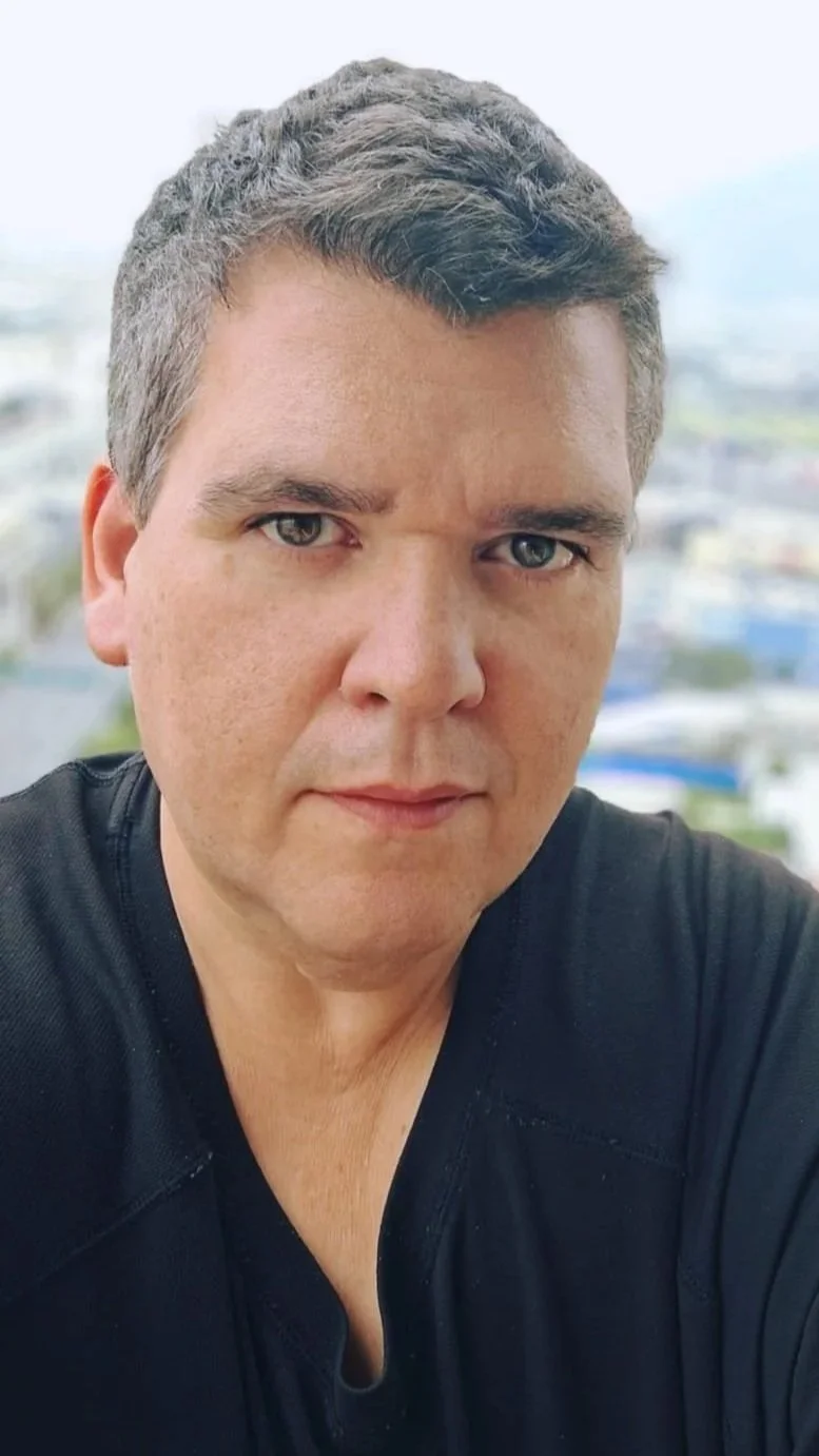 Retrato de un hombre con cabello castaño y ojos azules, usando camiseta negra, con fondo difuso de ciudad.