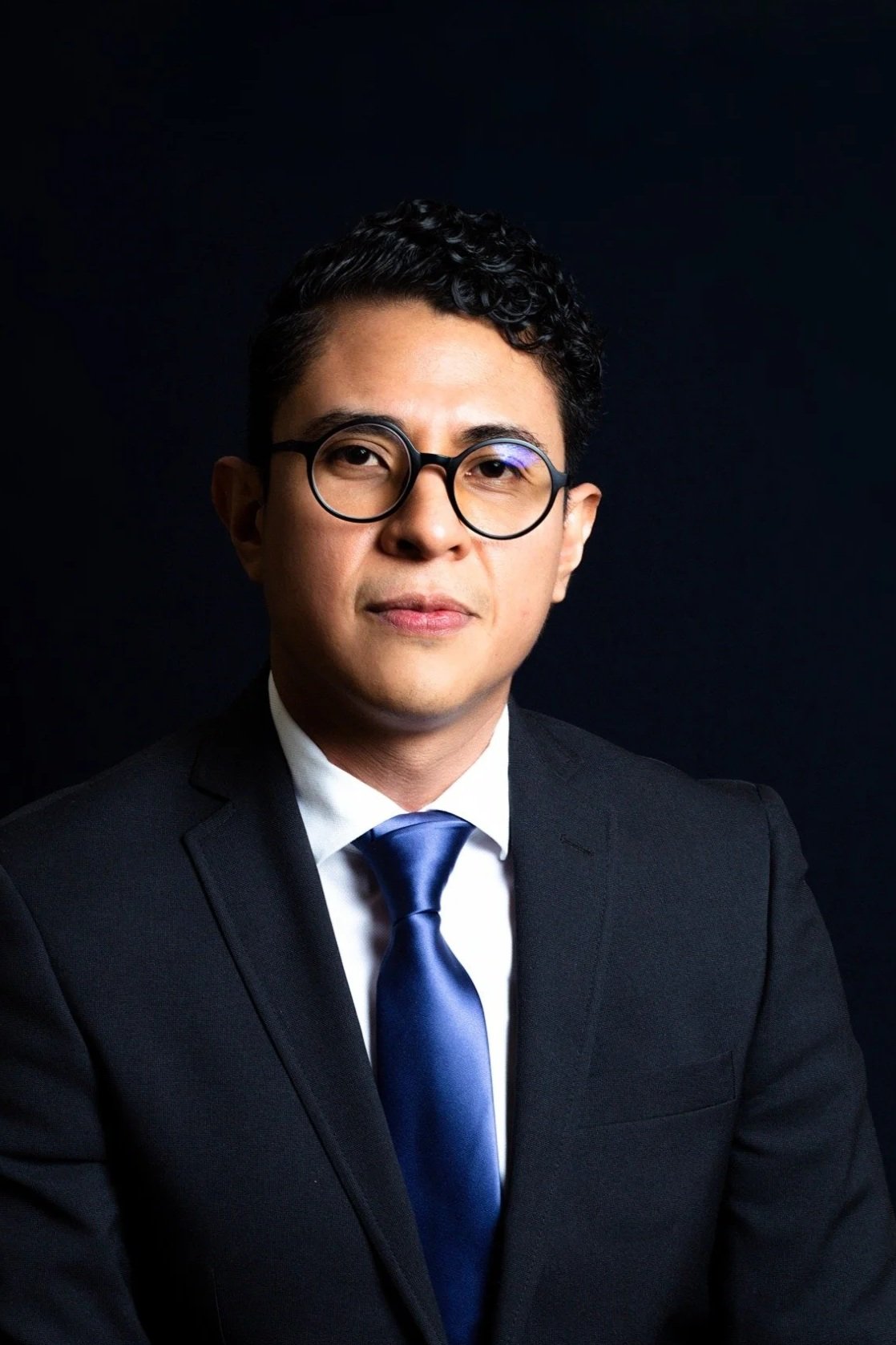 Hombre de cabello rizado, lleva gafas, vestido con saco negro, camisa blanca y corbata azul, fondo negro.