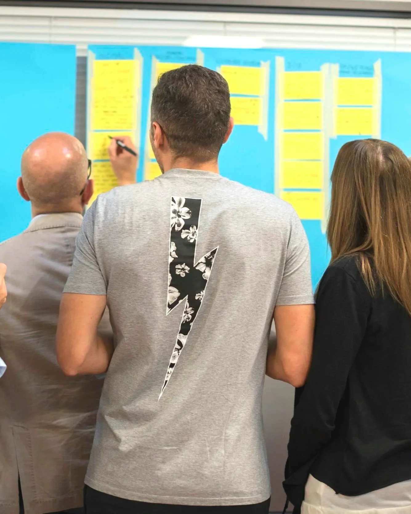 Tres personas observando un tablero con notas amarillas en una pared, en un entorno interior.