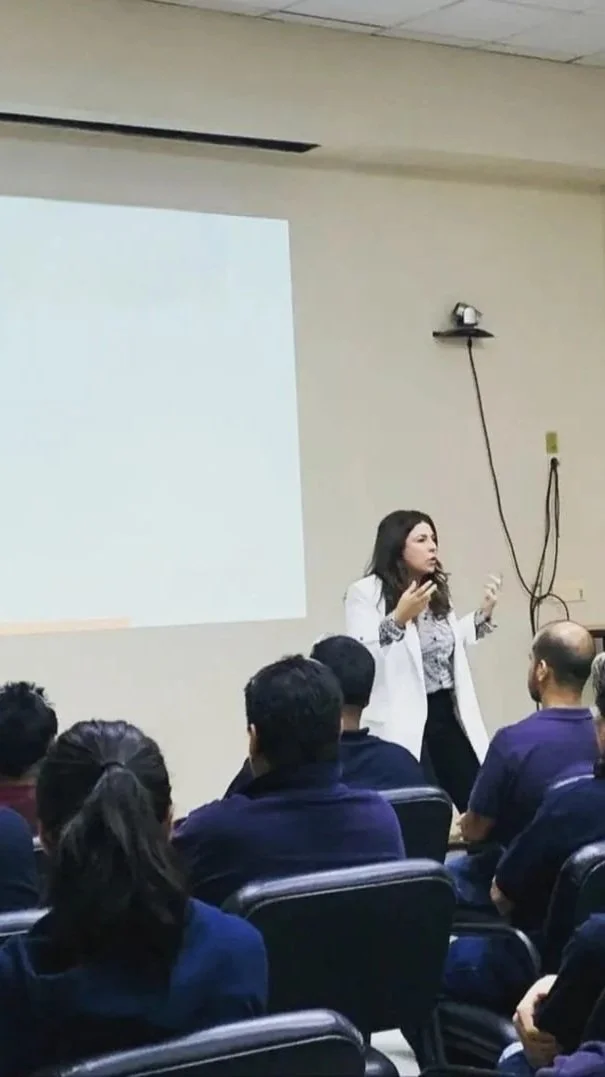 Mujer dando una charla o conferencia frente a una audiencia en una sala de reuniones o aula, con pantalla y proyector al fondo.