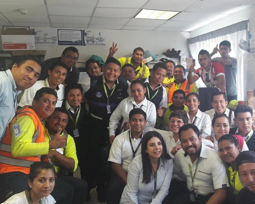 Grupo diverso de personas en una oficina, la mayoría con uniforme y algunos con chalecos de seguridad, algunos haciendo gestos con las manos y sonriendo.