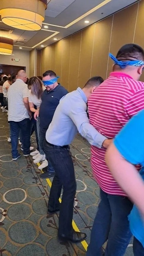 Personas en línea participando en un juego de soga ciega, con vendas en los ojos, en una sala con decoraciones en el techo y pared.