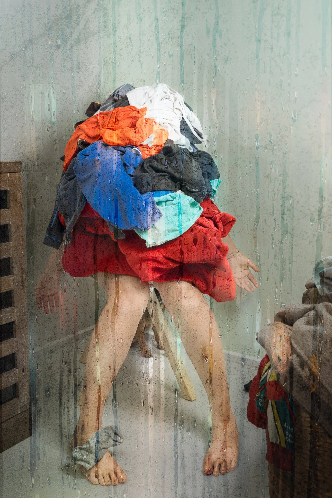 A statue of a person with multiple layers of colorful clothes piled on top of their head, seen through a rain-covered glass window.