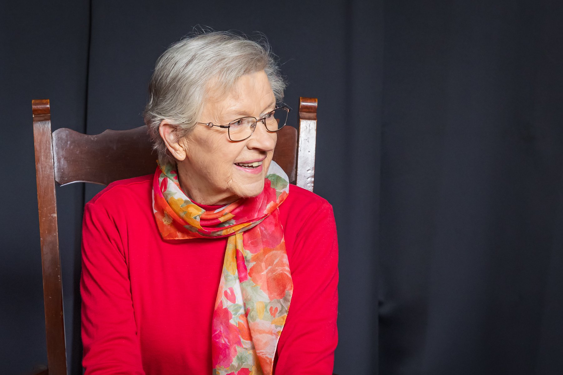 Elderly woman with glasses, wearing a bright scarf and red top, sitting on a wooden chair against a dark background.