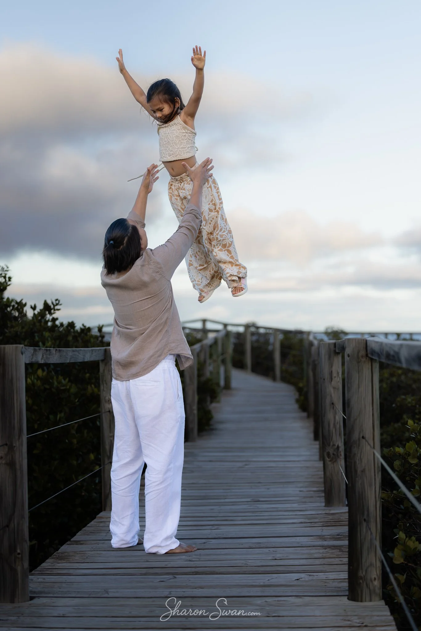 A woman is holding a young girl in the air on a wooden bridge outdoors at dusk, with clouds in the sky behind them.