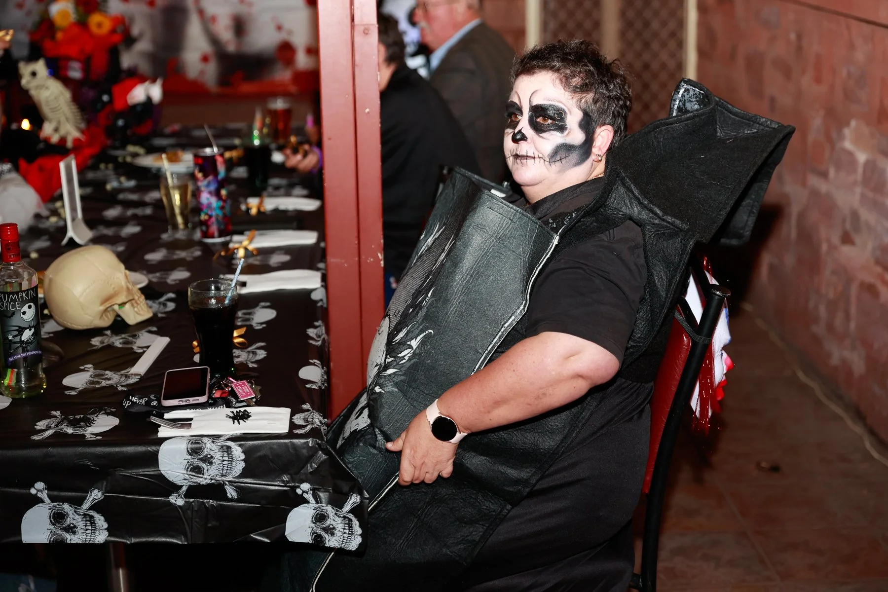 Person at Halloween party wearing skeleton face paint and costume, seated at a decorated table with skull-themed decorations.