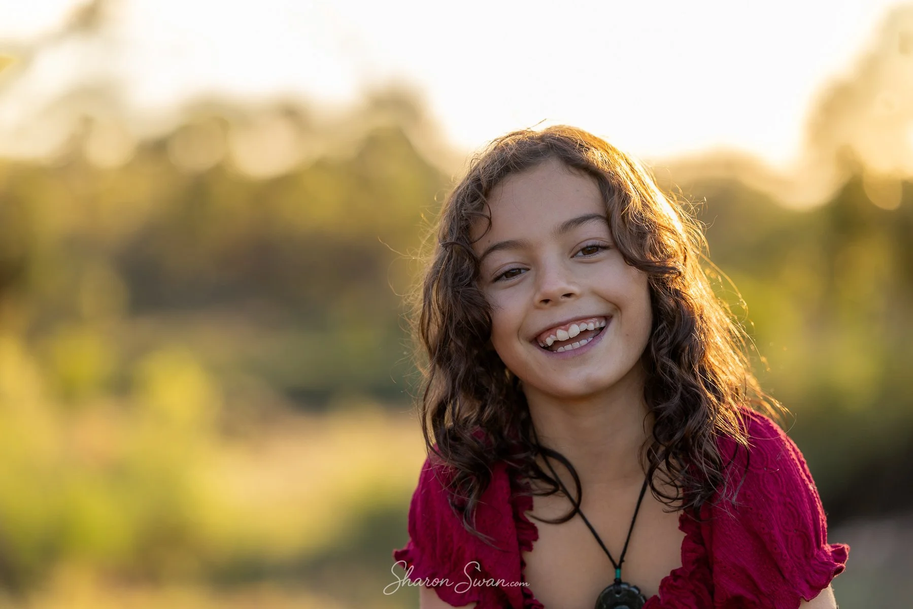 A young girl with long curly hair smiling outdoors during sunset, wearing a red top and a necklace.