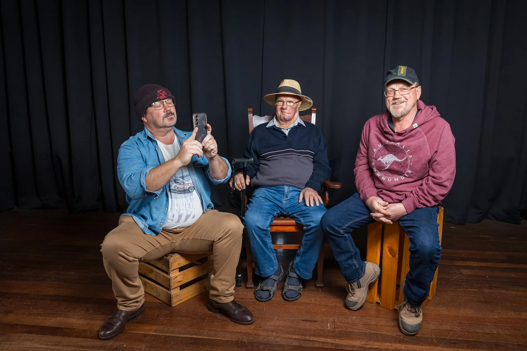 Three men sitting, one holding a smartphone, indoors with a black curtain backdrop.