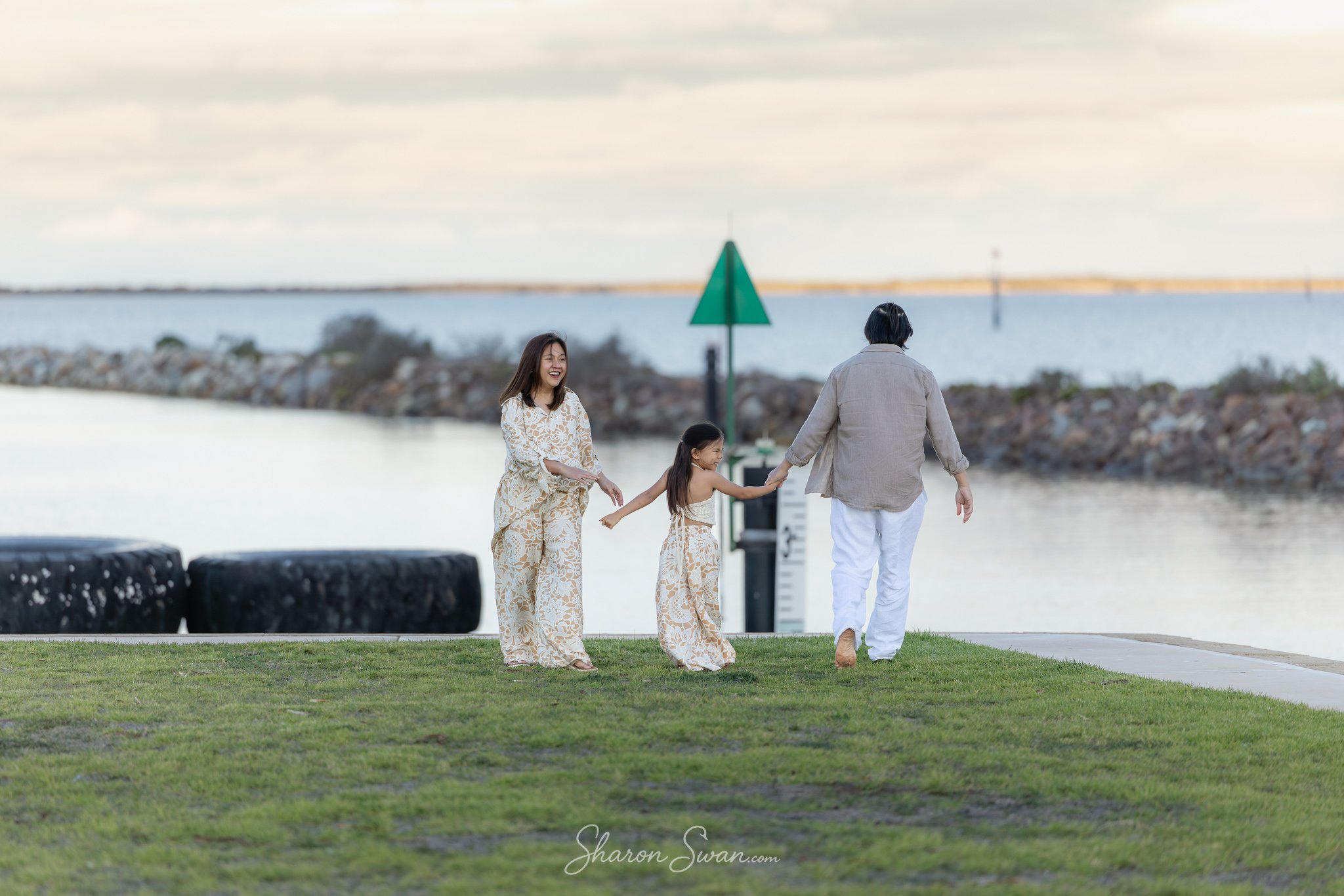 A family walking along a waterfront promenade with a woman, a man, and two young girls, all wearing light-colored, patterned clothing, with water and a cloudy sky in the background.