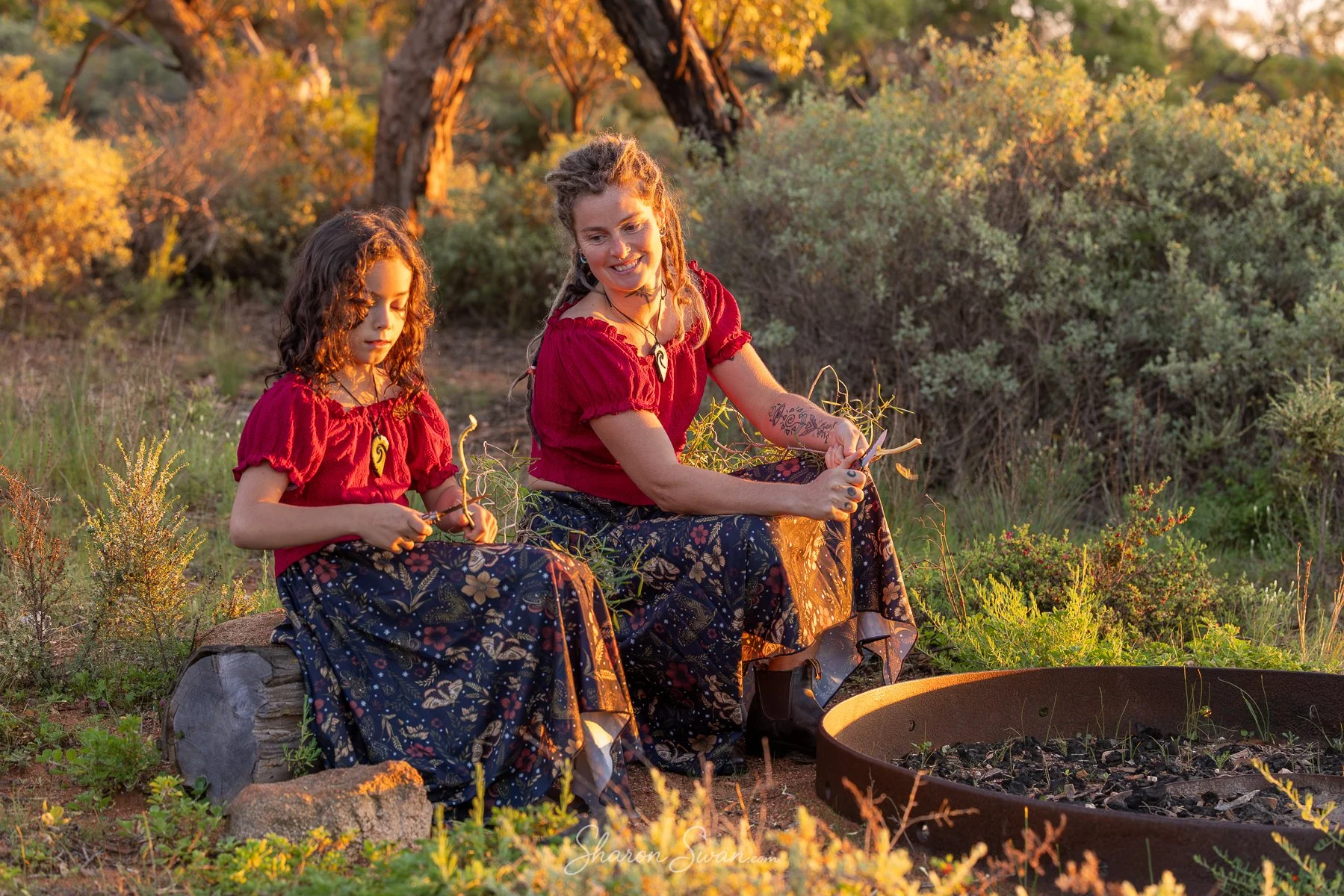A woman and a young girl sitting outdoors on a log, surrounded by greenery and trees with autumn foliage, both wearing matching red tops and dark patterned skirts, engaging in a nature activity during sunset.
