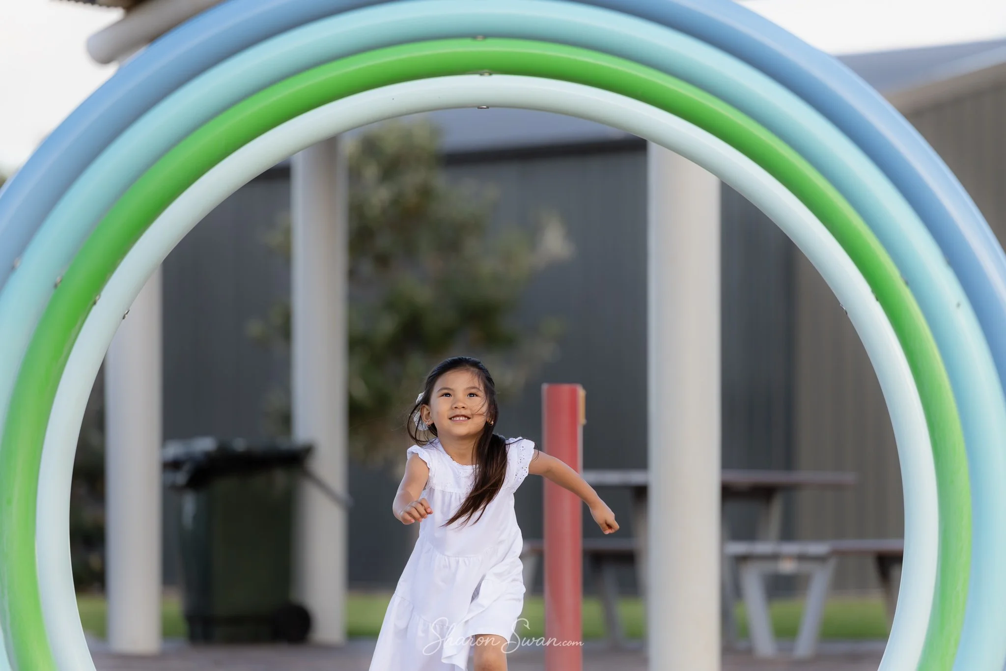 A young girl in a white dress running through a colorful playground with rainbow arches overhead.