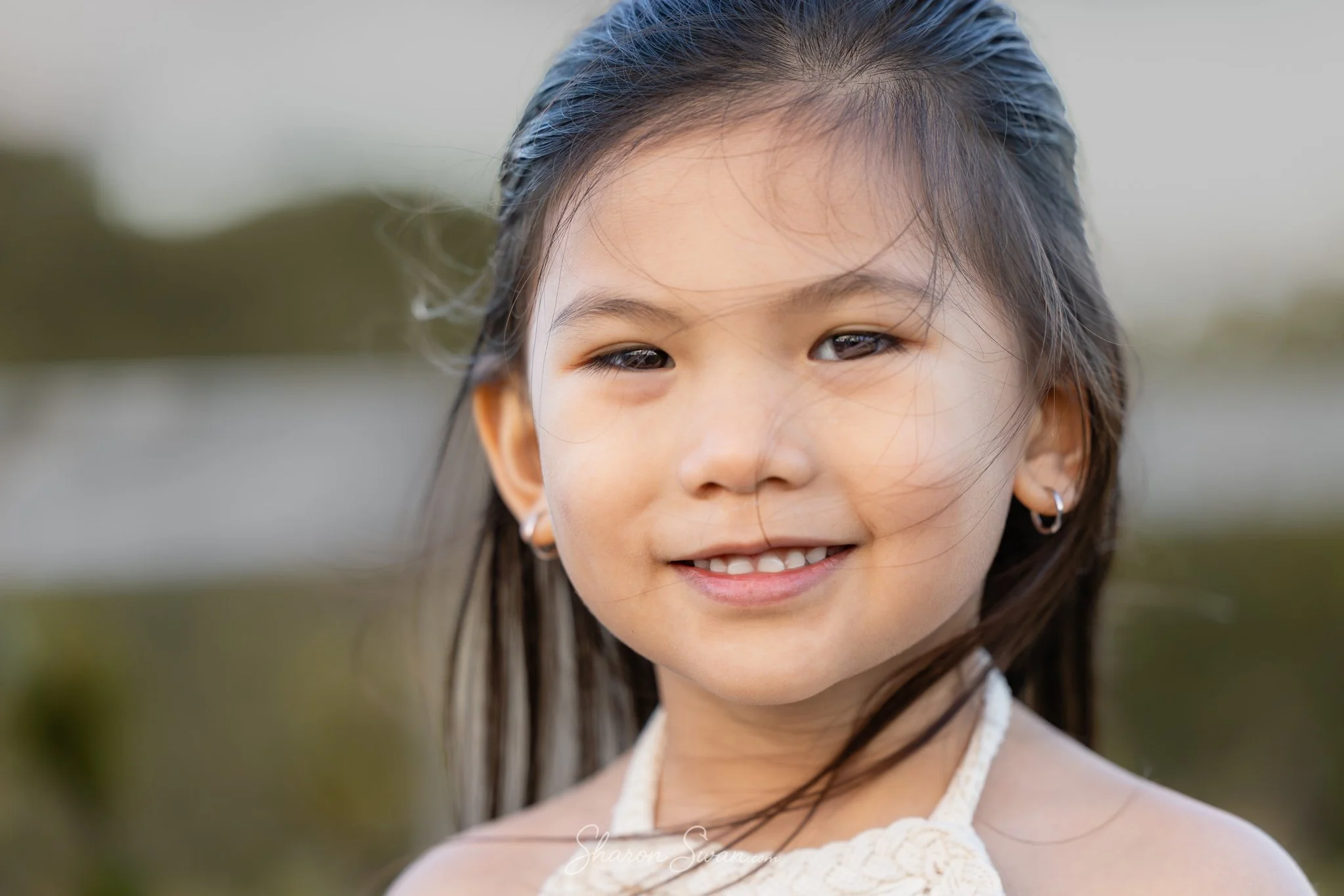 A young girl with long dark hair, smiling, wearing earrings and a white lace top outdoors.