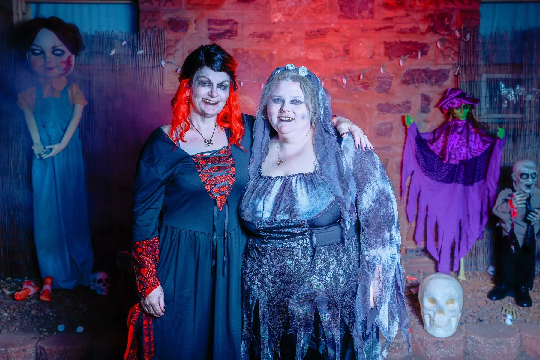 Two women in Halloween costumes standing in front of a haunted house-themed backdrop with a skeleton, a purple-clad figure, and a child mannequin in overalls.