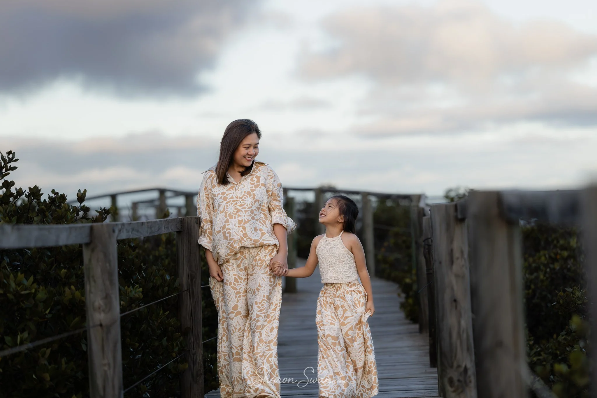 A woman and a young girl walking hand in hand on a wooden pathway outdoors, with a cloudy sky overhead. They are smiling at each other and wearing matching beige clothing with white floral patterns.