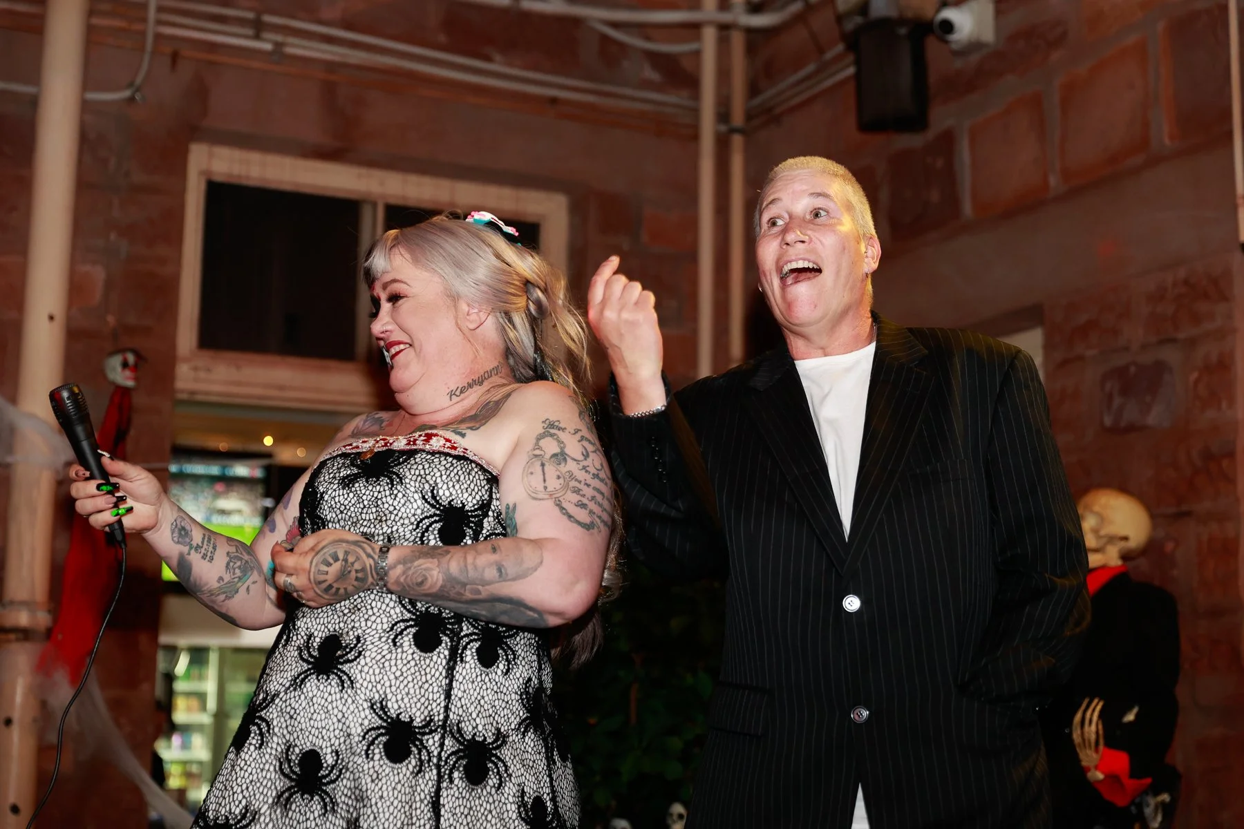 Two people laughing indoors; one holding a microphone. One is wearing a spider-patterned dress, the other a pinstripe suit. Halloween decorations like a skeleton are in the background.