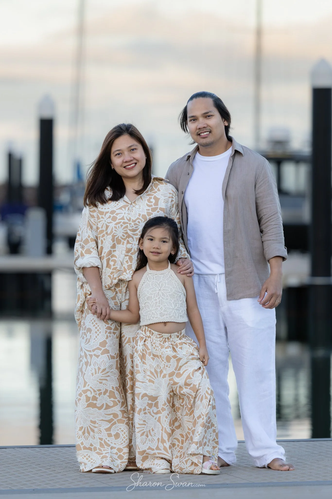 A family of four stands together outdoors near a body of water at sunset. The mother has long dark hair and is wearing a patterned beige dress. The father has short dark hair and is wearing a light gray shirt over a white t-shirt with white pants. Th