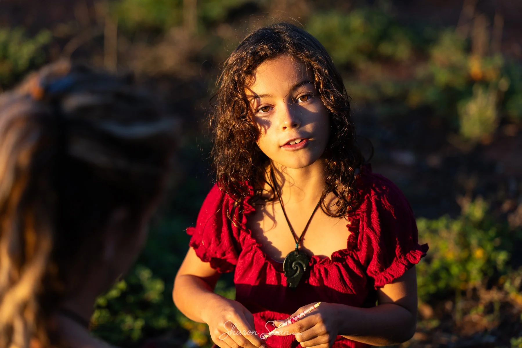 A young woman with curly hair and light brown eyes wearing a red ruffled top and a black spiral pendant necklace, standing outdoors during sunset.