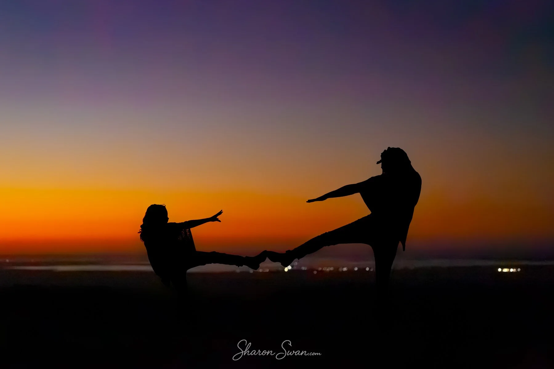 Silhouette of an adult and a child holding hands and balancing on a beach during sunset, with the sky transitioning from orange to purple.