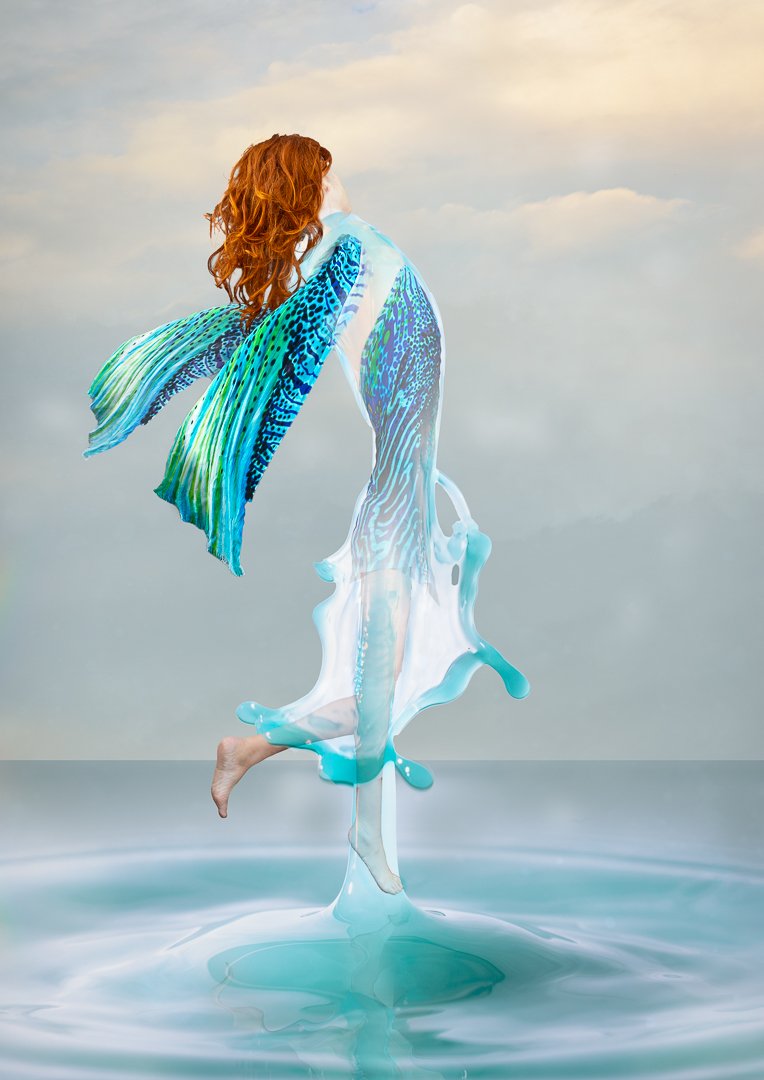 A person with red hair is surrounded by a surreal, digital flow of blue and green liquid resembling a mermaid's tail and fins, set against a cloudy sky background.