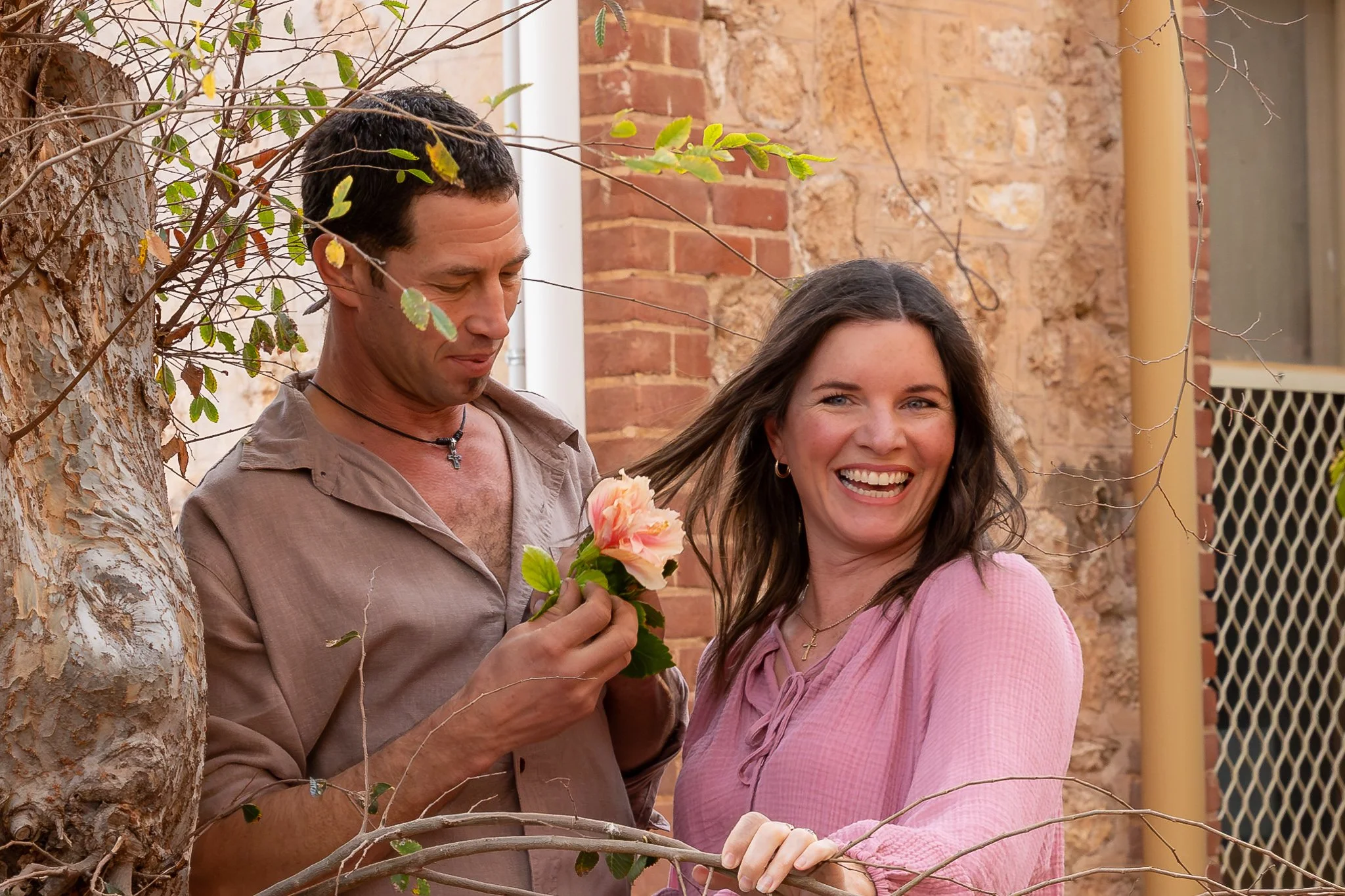 A man and woman outdoors, man holding a peach-colored flower, woman smiling, with a brick wall and tree branches in the background.
