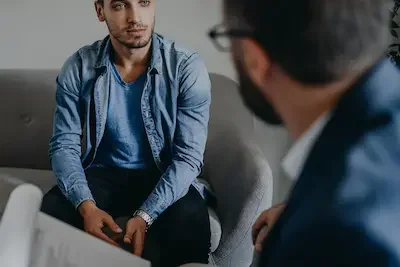 An image of an adult male sitting on a couch in conversation with a therpaist in rehab