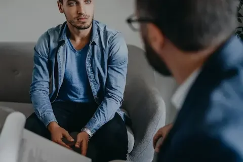 Image of an adult male sitting on a couch talking to his therapist in rehab