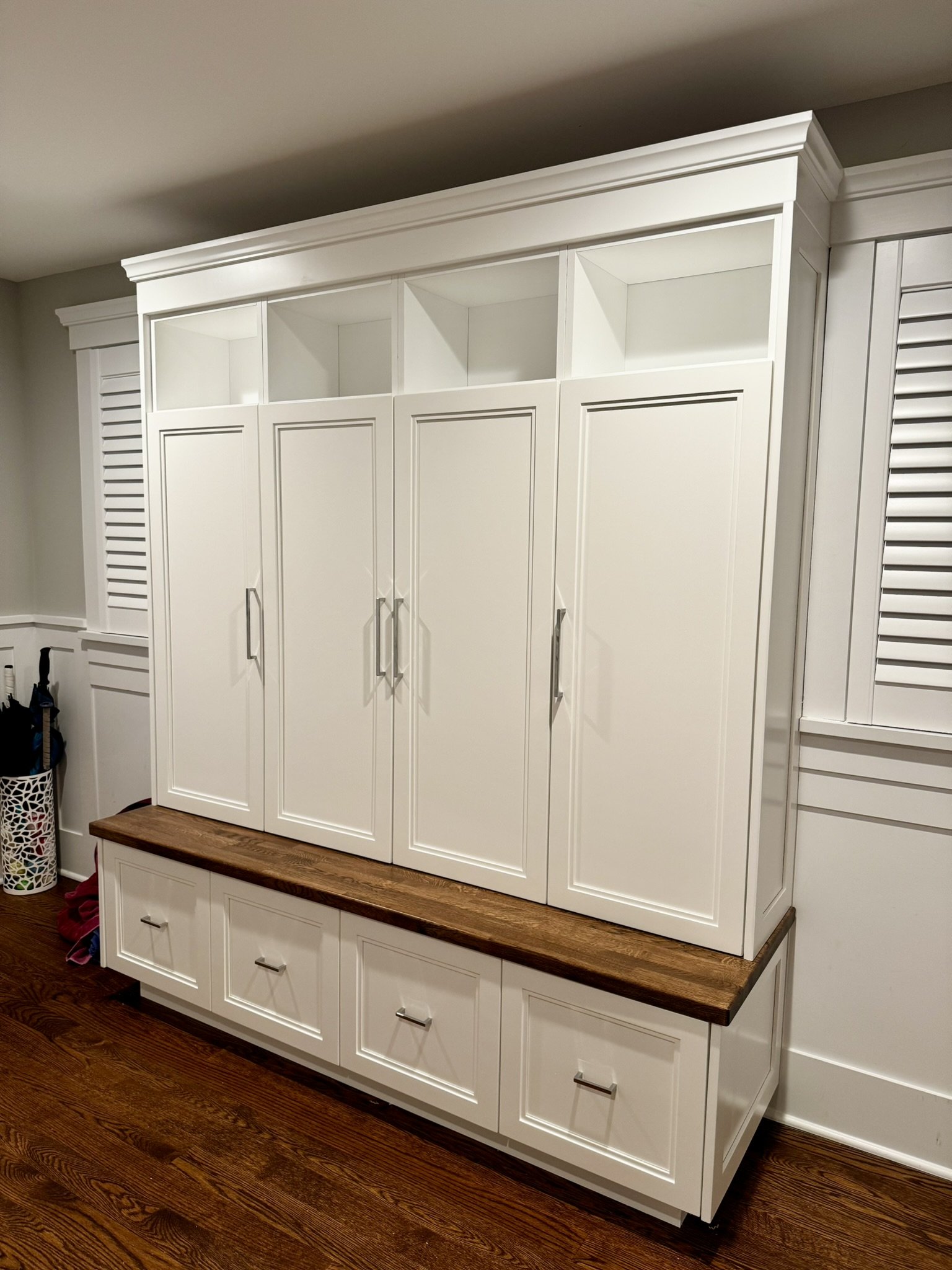 White built-in cabinet with four central doors, open shelves above, and drawers below, with a wooden top, in a room with hardwood floors.
