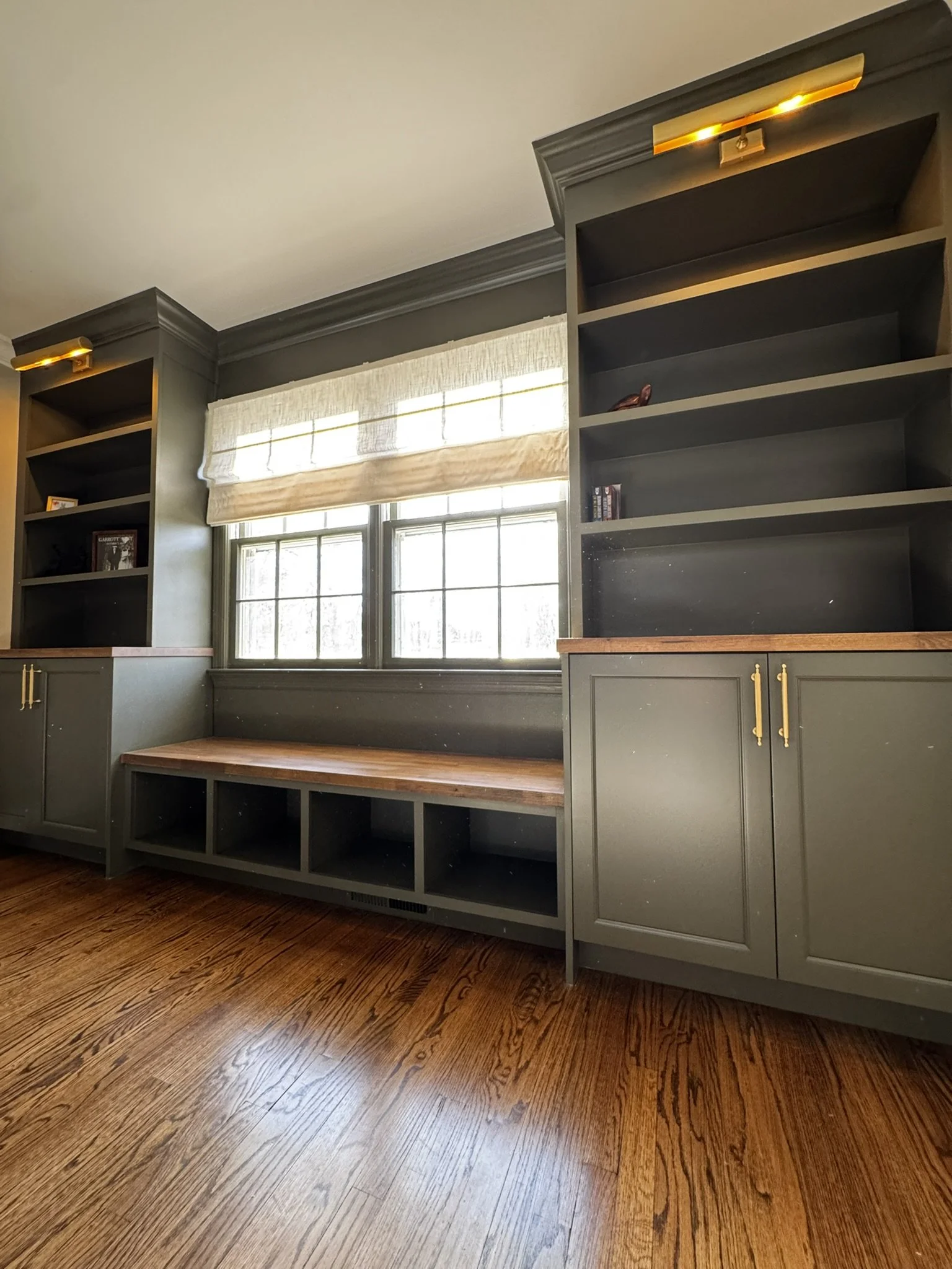 Built-in window seat with storage and tall green bookshelves on each side, a window with a beige roman shade, and hardwood floors.