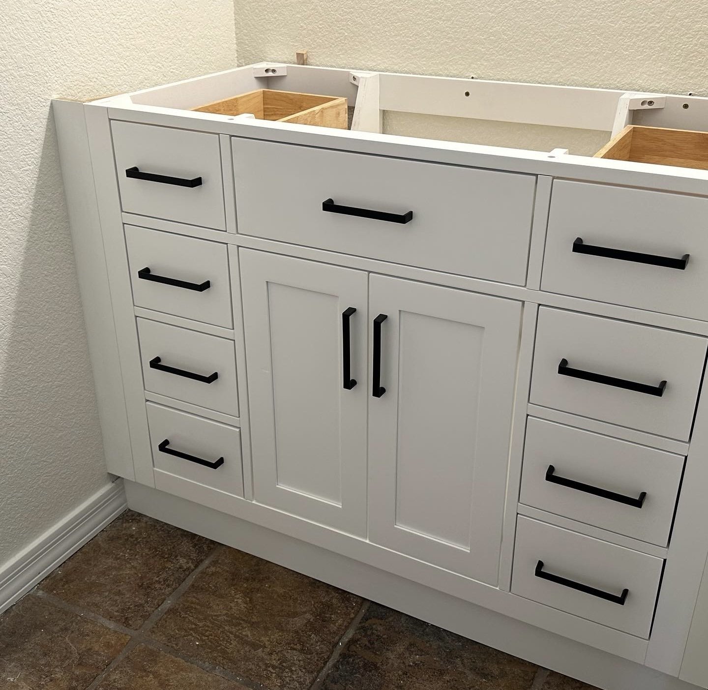 White kitchen cabinet with black handles under construction, against a beige wall, on a tiled floor.