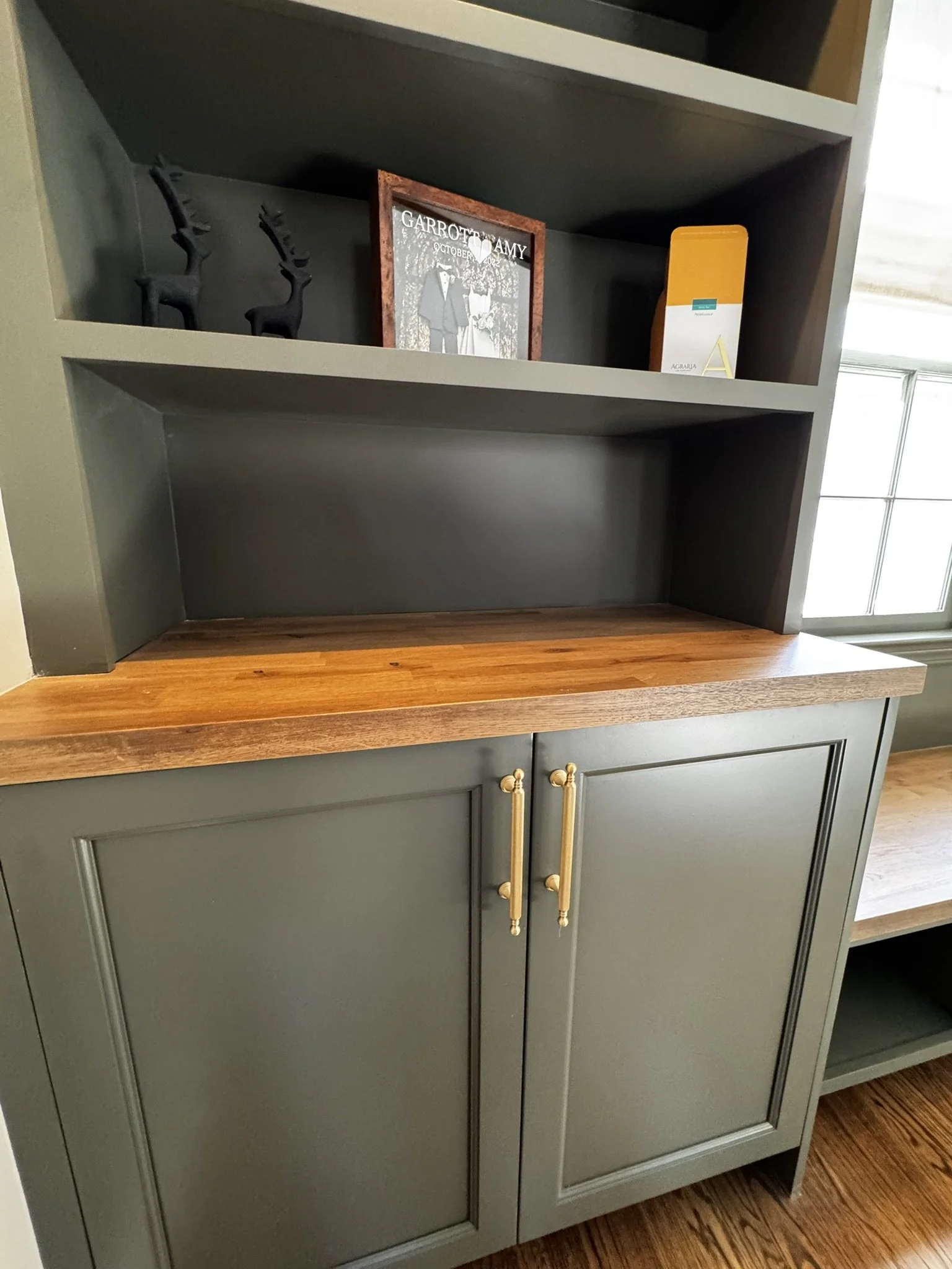 A storage cabinet with a wooden countertop, green cabinet doors with gold handles, and open shelves above decorated with small black deer figurines, a framed artwork, and a yellow and white box. There is a window to the right letting in natural light