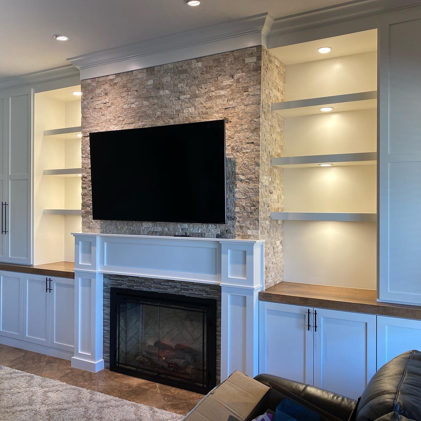 Living room with fireplace, mounted TV on a stone accent wall, white built-in cabinets, and empty shelves with built-in lighting.