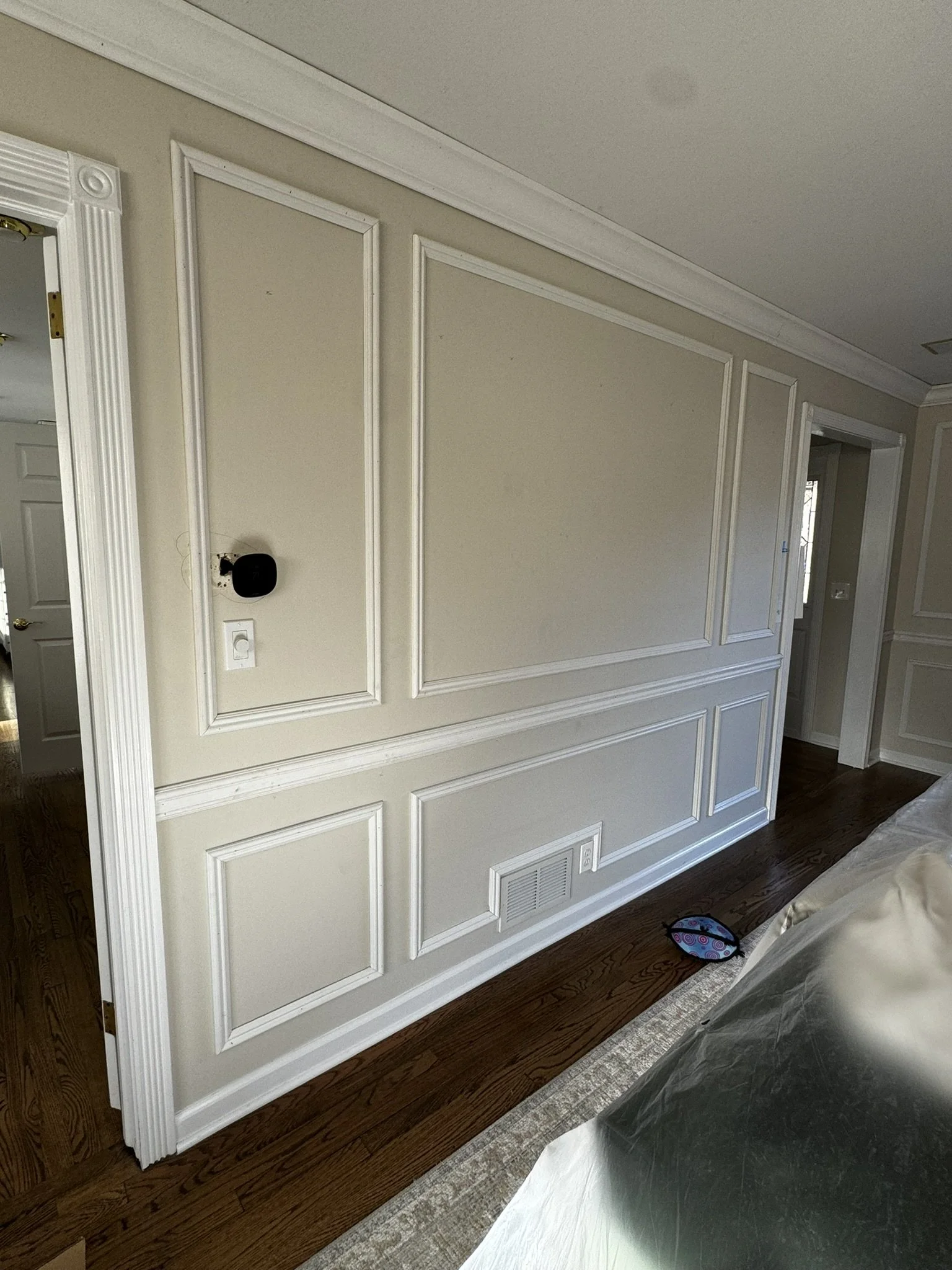 Empty living room wall with decorative molding, a black security camera, and a white light switch, with hardwood flooring and part of a bed covered in plastic visible in the foreground.