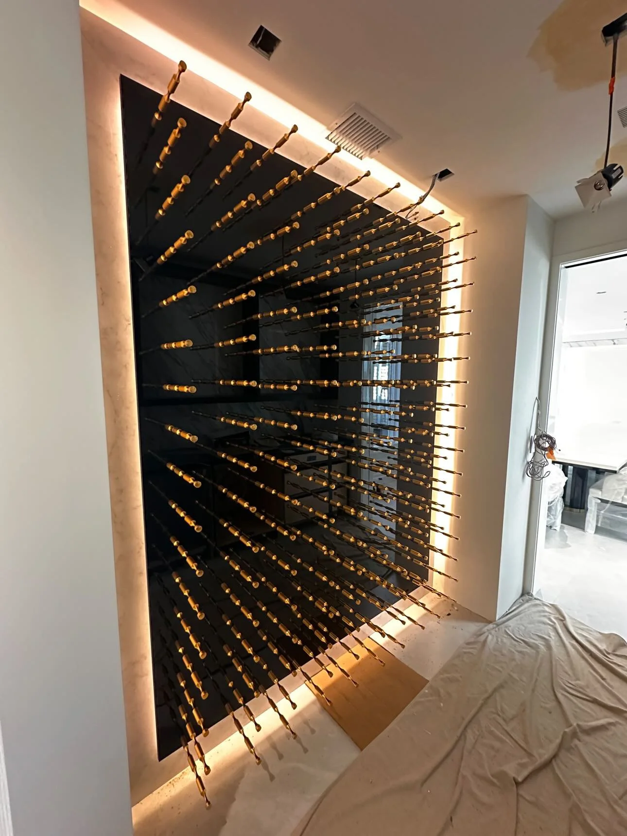 A wall-mounted art piece featuring numerous black rods with yellow caps, arranged in a radial pattern on a black background, with backlighting creating a glowing effect.